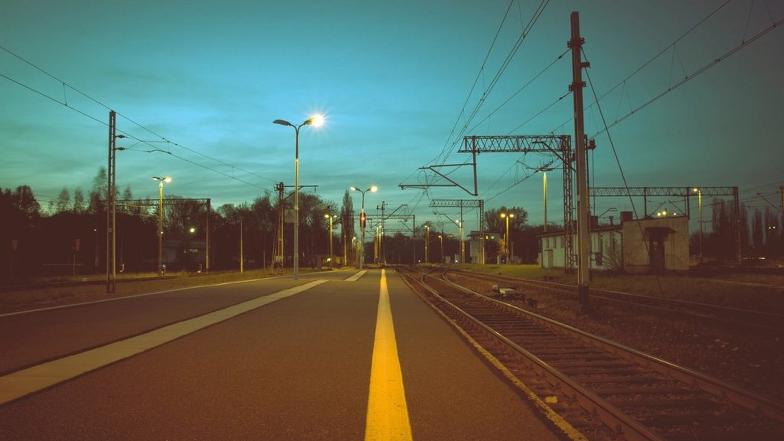 夕暮れの駅のホームに黄色い線がまっすぐ伸び、架線の柱と街灯が並ぶ写真